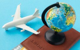 Passaporte de couro marrom com um pequeno globo terrestre em cima, acompanhado de duas passagens aéreas de primeira classe e uma miniatura de avião branco, organizados sobre um fundo azul claro.