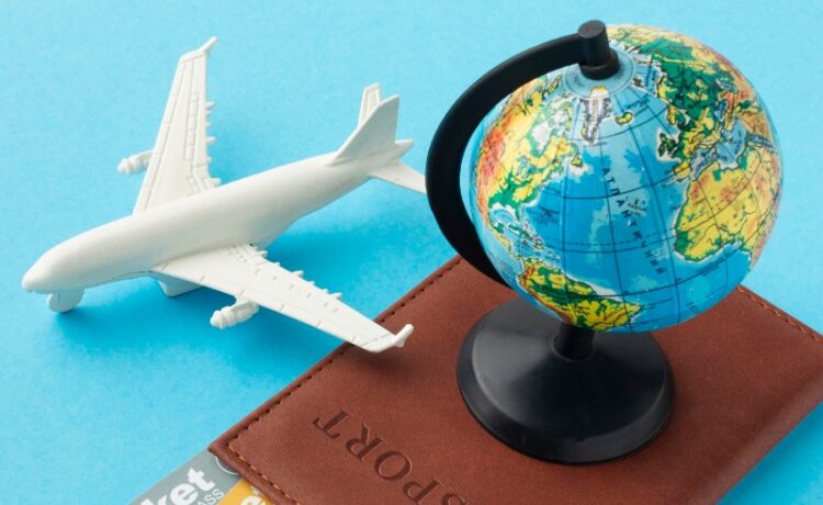 Passaporte de couro marrom com um pequeno globo terrestre em cima, acompanhado de duas passagens aéreas de primeira classe e uma miniatura de avião branco, organizados sobre um fundo azul claro.