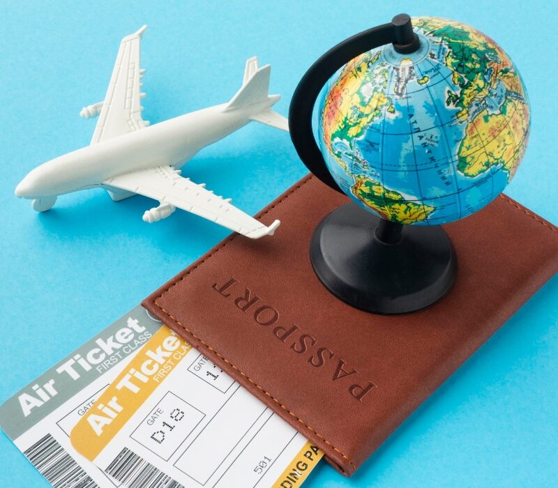 Passaporte de couro marrom com um pequeno globo terrestre em cima, acompanhado de duas passagens aéreas de primeira classe e uma miniatura de avião branco, organizados sobre um fundo azul claro.