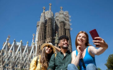 Três pessoas fazem selfie diante de basílica em imagem sobre brasileiros na Espanha
